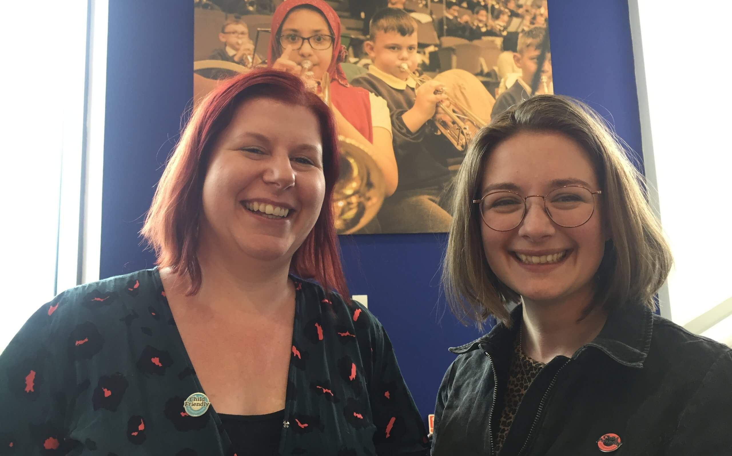 New SCEP team members: Jodie Sadler on the left is a white woman with red hair and Frankie McCormick on the right has brown hair and glasses. They are both smiling and standing in front on photograph of children playing musical instruments. 