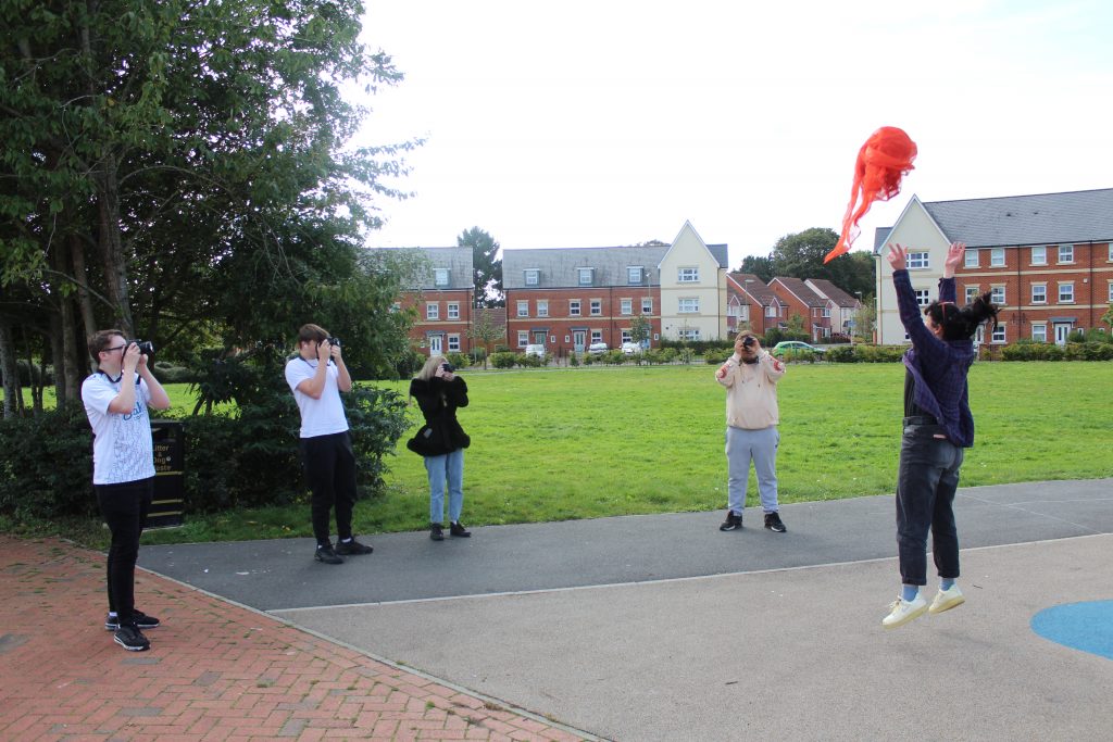 A group of five young people are stood outside. One of them is jumping and throwing a red object in the air, while the other four are holding digital cameras and taking photos of the object. 