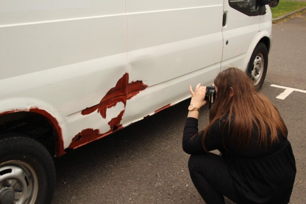 A person with long hair crouches down to take an artistic photo of the rust on a white van