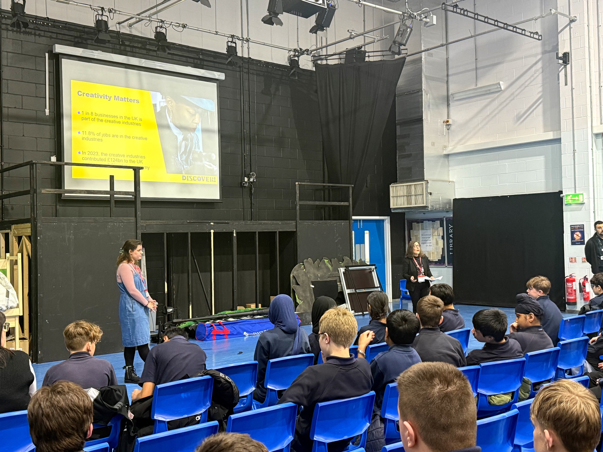 Philippa and Lucy present an assembly, a few rows of students are visible.