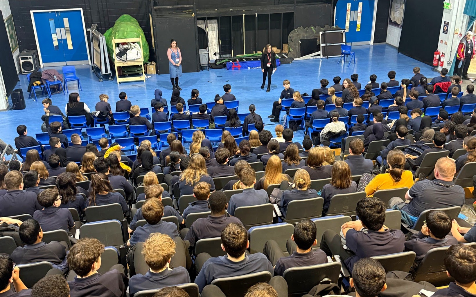 Philippa and Lucy presenting an assembly in a school hall, the audience of children are visible.